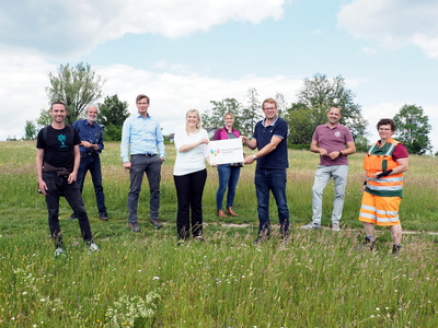 8 Personen in einer Blumenwiese, die vorderen zwei Personen halten ein Schild mit der Aufschrift Bienenfreundliche Gemeinde